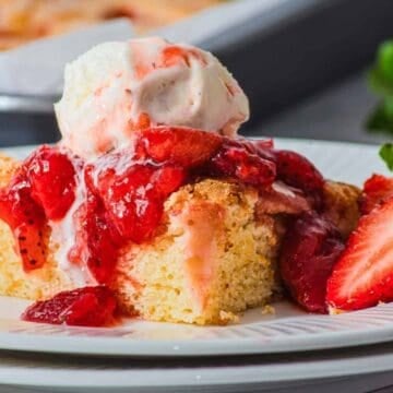 A square piece of yellow cake topped with strawberry sauce and a scoop of vanilla ice cream is on a white plate, with a fresh strawberry and a cake pan in the background.