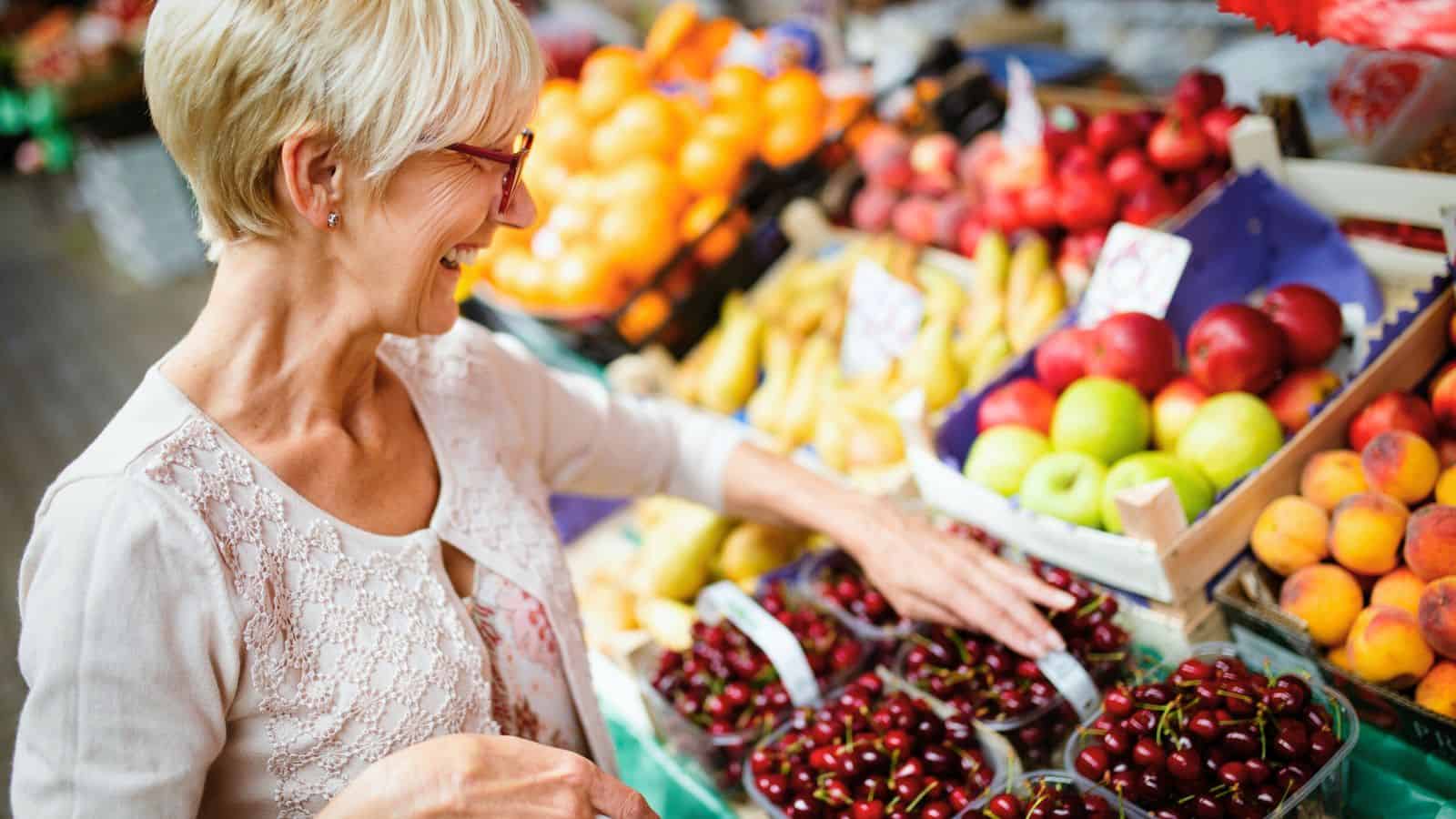 An older woman with short blonde hair and glasses smiles while selecting cherries from a display of assorted fruits at an outdoor market. Apples, peaches, bananas, and oranges are visible nearby.