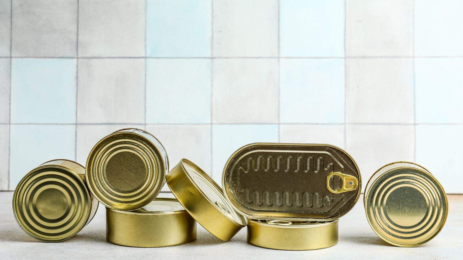 Several closed gold-colored tin cans and one opened rectangular can are arranged on a light surface in front of a tiled wall. The cans are grouped together, some standing and some lying down.