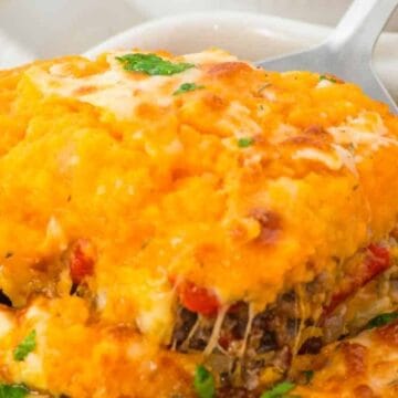 A close-up view of a casserole dish containing shepherd's pie topped with golden mashed sweet potatoes and melted cheese, garnished with chopped herbs and served with a spoon.