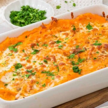 A white baking dish filled with baked sweet potato casserole topped with melted cheese and chopped herbs, placed on a wooden board with bowls of fresh herbs in the background.