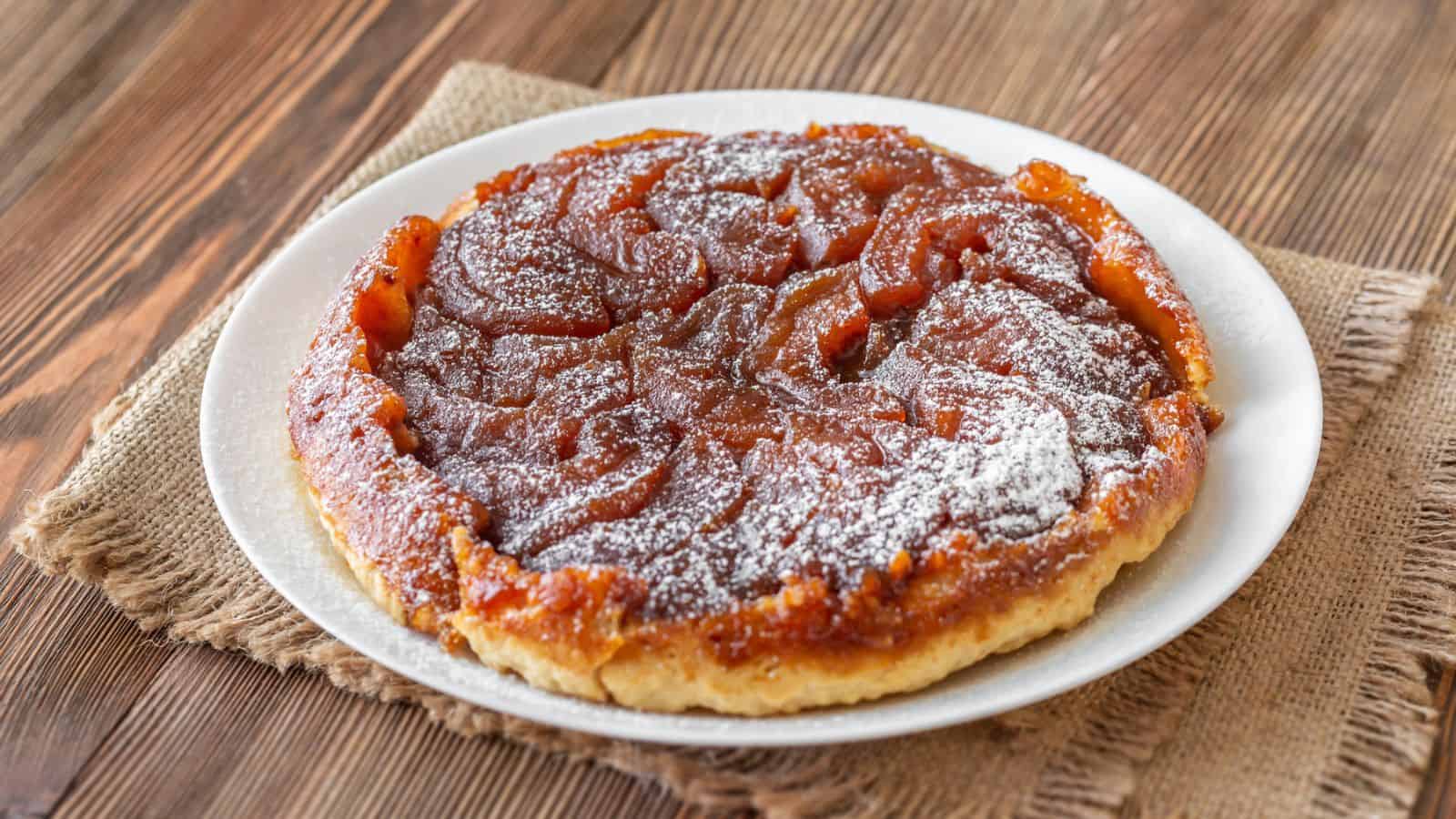A tarte Tatin sits on a white plate atop a piece of burlap on a wooden table. The caramelized apple topping is visible and dusted lightly with powdered sugar.
