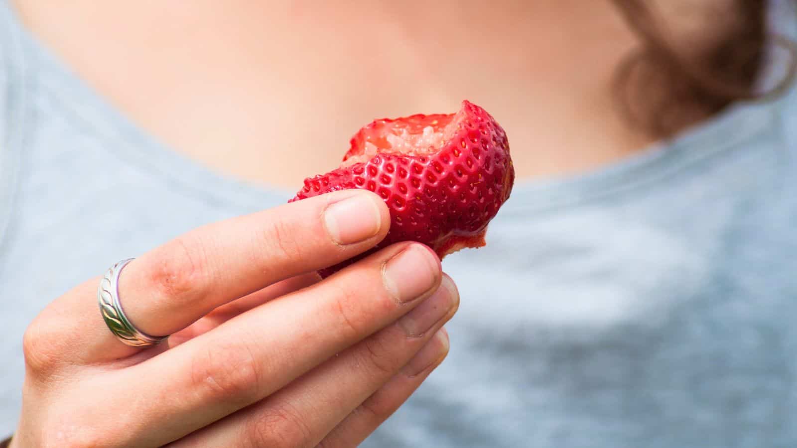 A person wearing a light gray shirt holds a partially eaten strawberry between their fingers. The person has a silver ring on their index finger and their face is not visible.