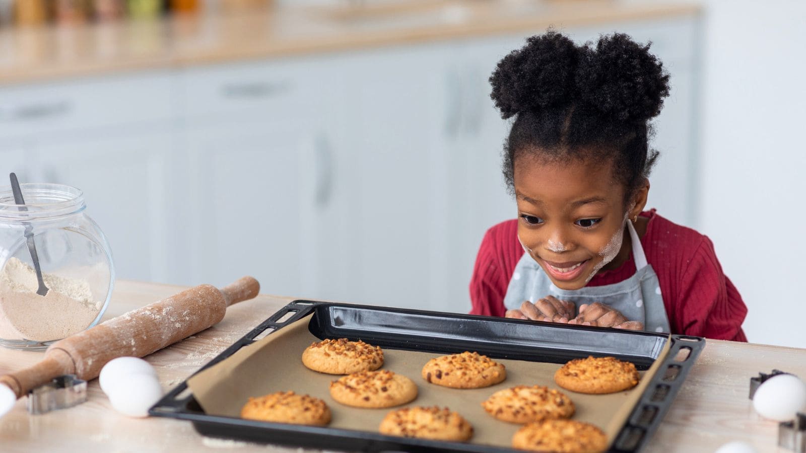 A young child wearing an apron smiles and looks closely at a tray of freshly baked cookies on a kitchen counter. A rolling pin, flour jar, and eggs are also on the counter.