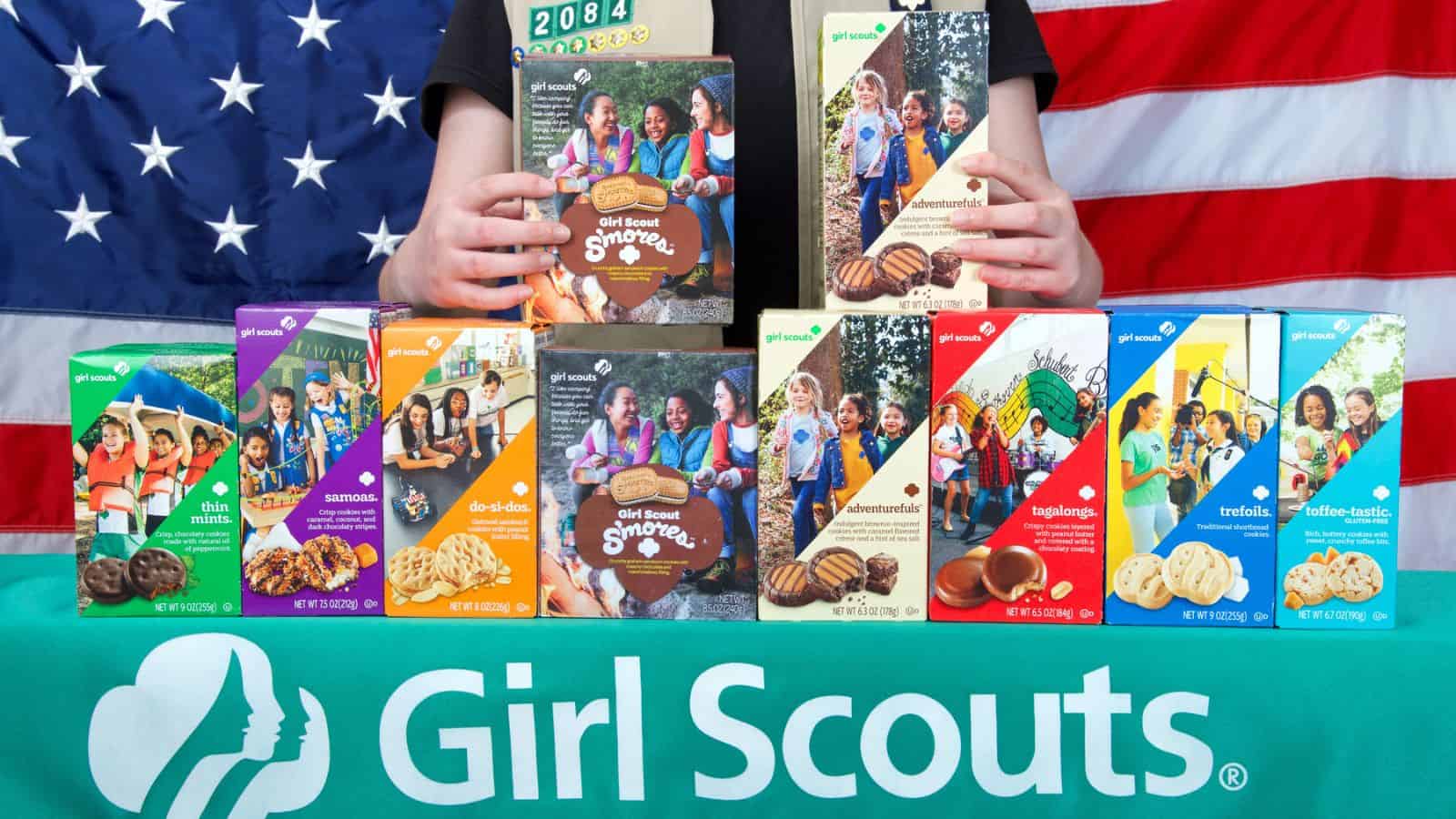 A person stands behind a table with nine varieties of Girl Scout cookies displayed in boxes. The table has a Girl Scouts logo, and an American flag hangs in the background.