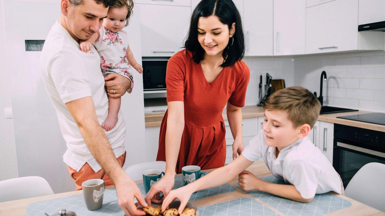 A man holding a young child, a woman, and a boy are standing around a kitchen table, reaching for cookies on a plate. There are also two mugs on the table. The setting is a modern kitchen.