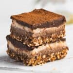Two Tiramisu Protein Bars are stacked on top of each other. Each bar has a base layer of oats and nuts, a creamy middle layer, and a cocoa-dusted top. The bars rest on white parchment paper, showcasing their rich, dessert-inspired appeal.