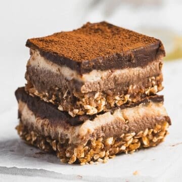 Two Tiramisu Protein Bars are stacked on top of each other. Each bar has a base layer of oats and nuts, a creamy middle layer, and a cocoa-dusted top. The bars rest on white parchment paper, showcasing their rich, dessert-inspired appeal.