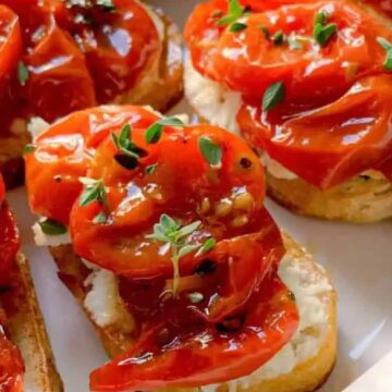 Slices of toasted bread topped with creamy cheese, roasted tomatoes, and fresh green herbs are arranged on a white plate in close-up.