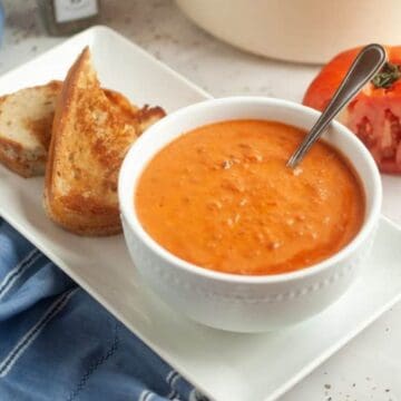 A white bowl of tomato soup with a spoon sits on a rectangular plate next to two slices of toasted bread. A halved tomato and a blue cloth are nearby on a white surface.