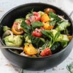 A black bowl filled with a Tomato and Smashed Cucumber Salad made of chopped tomatoes, cucumbers, onions, and fresh herbs, with a spoon resting inside the bowl.