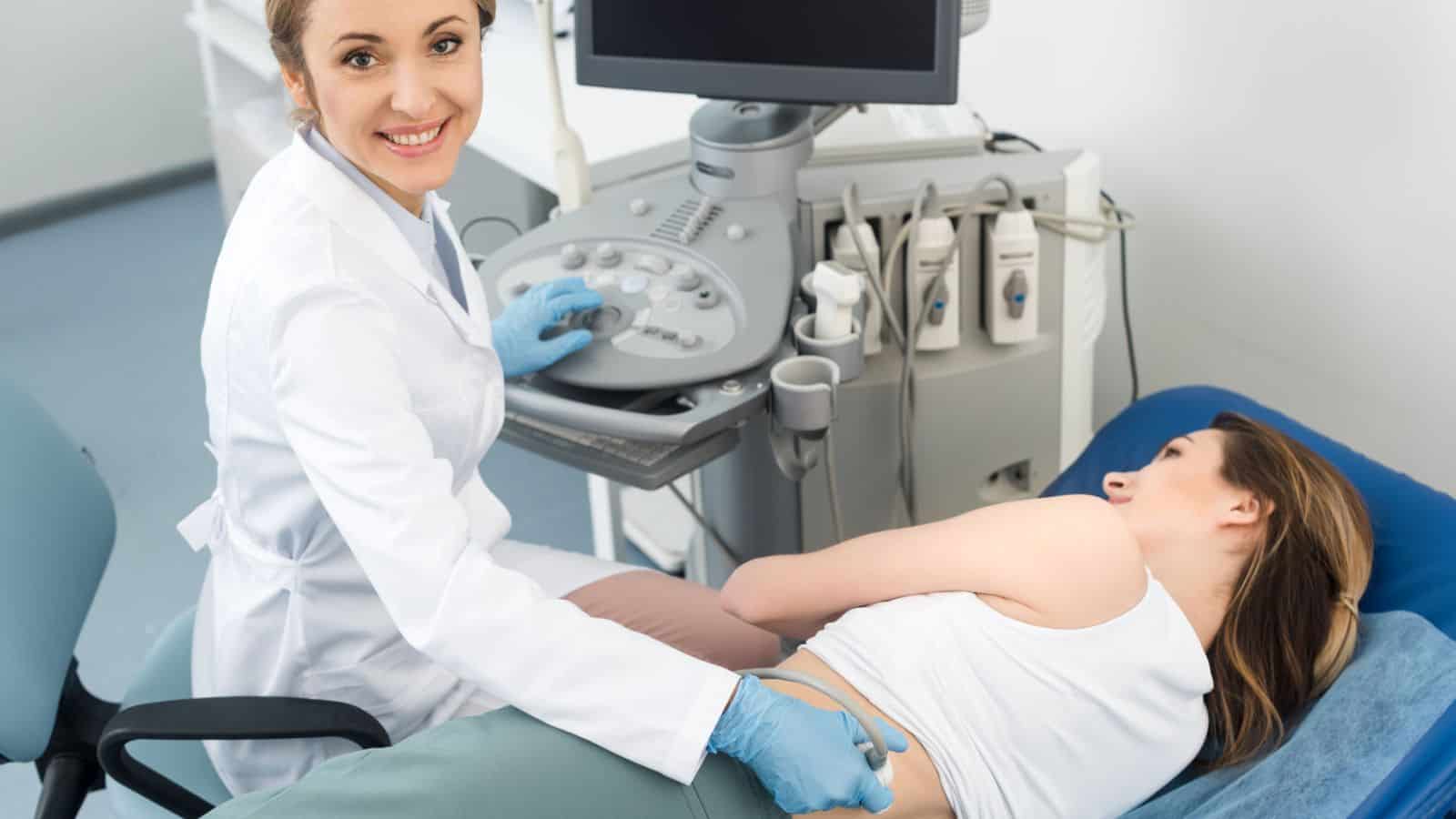 A healthcare professional performs an ultrasound scan on a patient lying on an examination table while looking at the camera and smiling. The patient is lying on their side with an exposed abdomen.