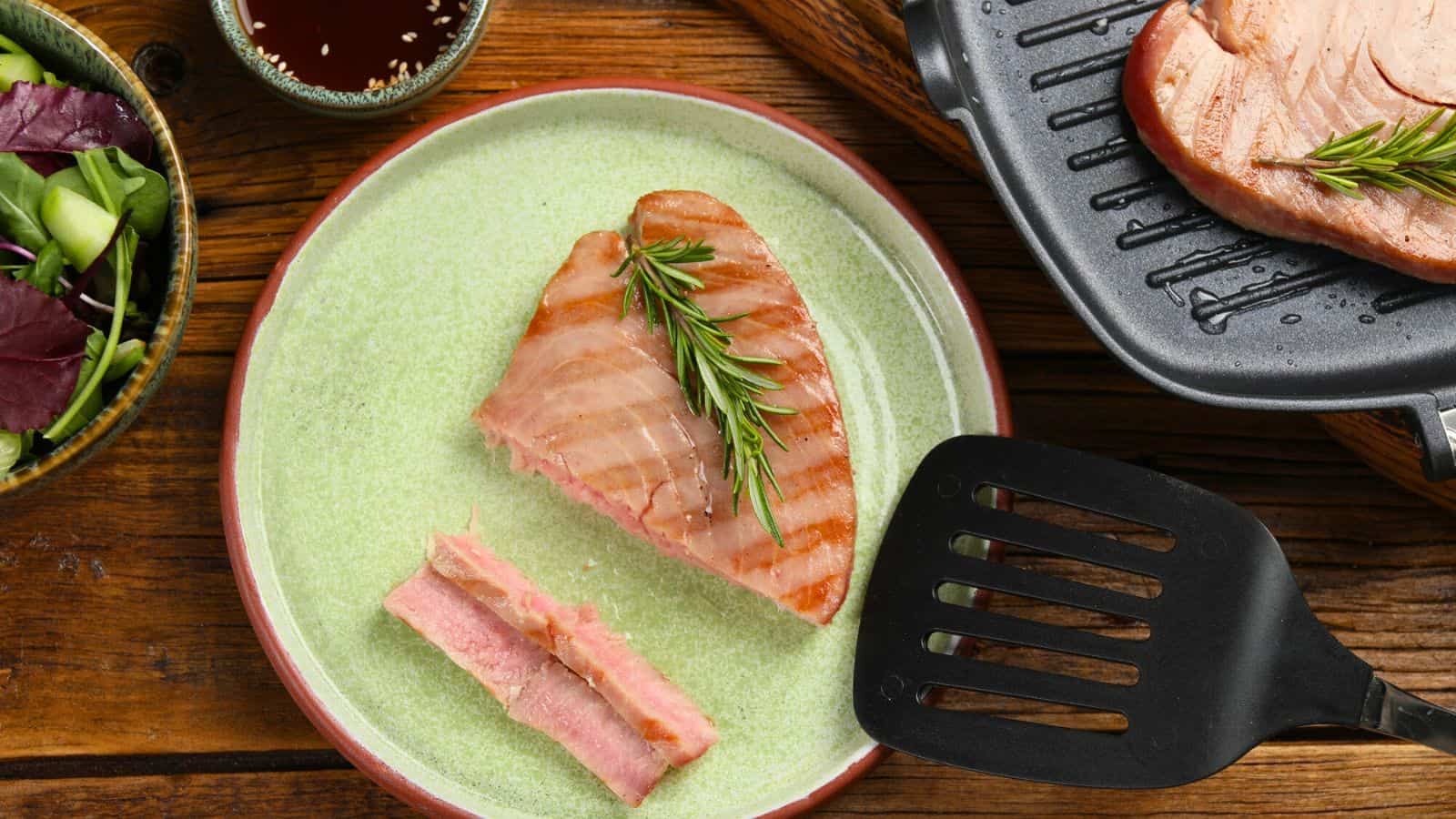A cooked tuna steak topped with a sprig of rosemary is on a plate next to two sliced pieces. A spatula, another steak on a grill pan, a bowl of greens, and a small bowl of sauce are nearby on a wooden surface.