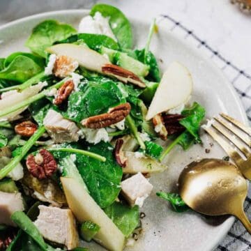 A plate of salad with spinach, slices of pear, pecans, chunks of chicken, cucumber, and cheese sits on a white surface next to a gold fork and spoon on a checkered napkin.