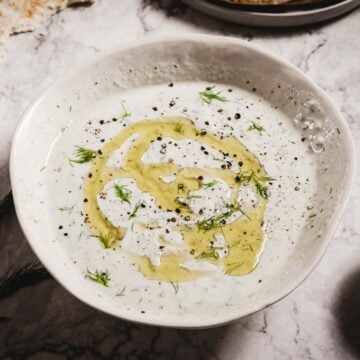 A white bowl filled with creamy Tzatziki Sauce, garnished with olive oil, fresh dill, and cracked black pepper, placed on a marble surface.