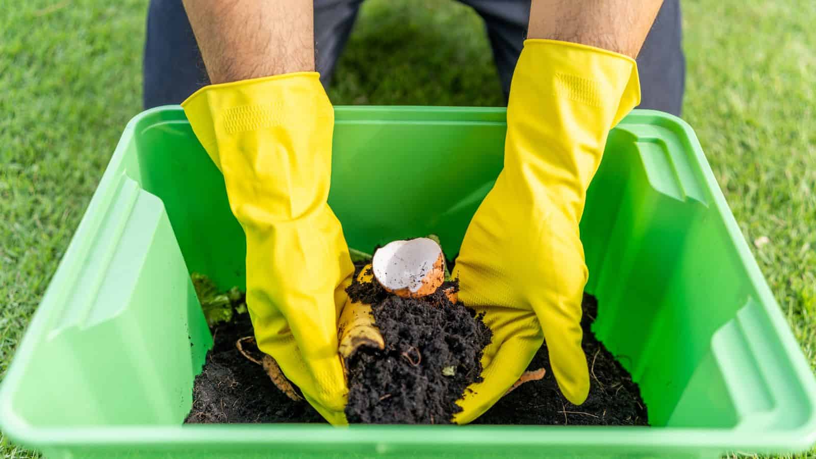 A person wearing yellow rubber gloves holds compost, including soil, a banana peel, and an eggshell, inside a green plastic container on grass.