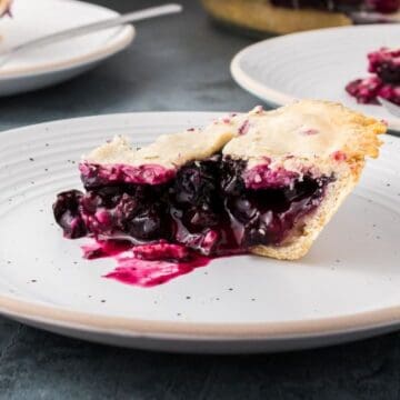 A slice of blueberry pie with a golden crust and purple filling sits on a white plate, with another slice and a pie dish in the background. Some blueberry filling has leaked onto the plate.