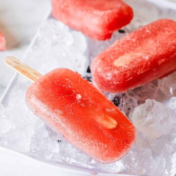 Three red popsicles on wooden sticks rest on a tray filled with ice cubes. A slice of watermelon and a lemon wedge are visible in the background on a white surface.