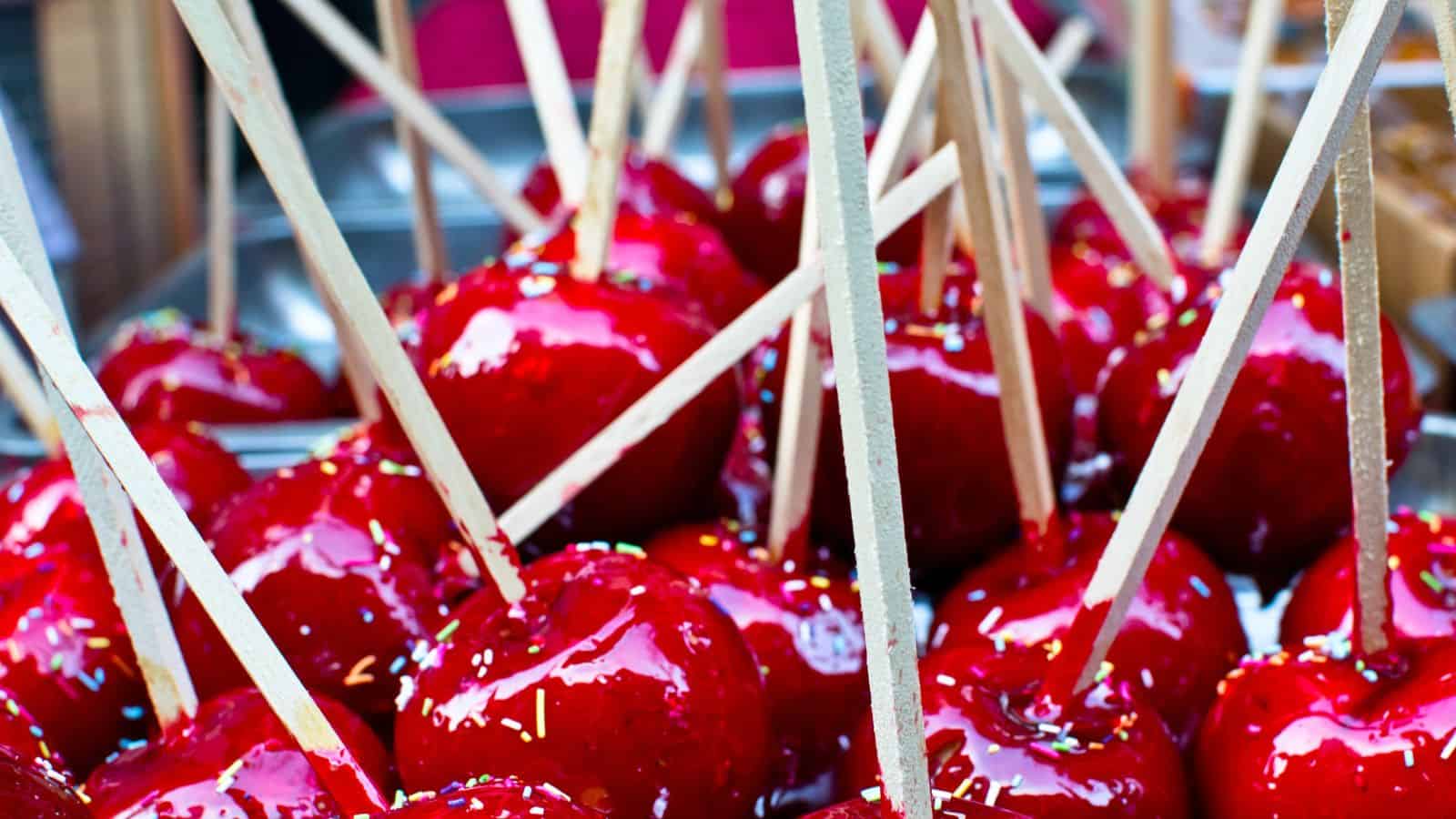 Several bright red candy apples on wooden sticks are grouped together, their glossy surfaces reflecting light. The apples are coated in a shiny candy glaze, and some have small colorful sprinkles.