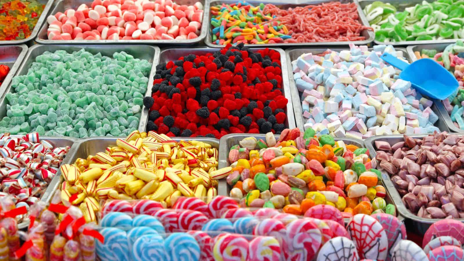 Assorted colorful candies displayed in metal trays, including gummies, marshmallows, hard candies, and lollipops, with a blue plastic scoop resting on one of the trays.
