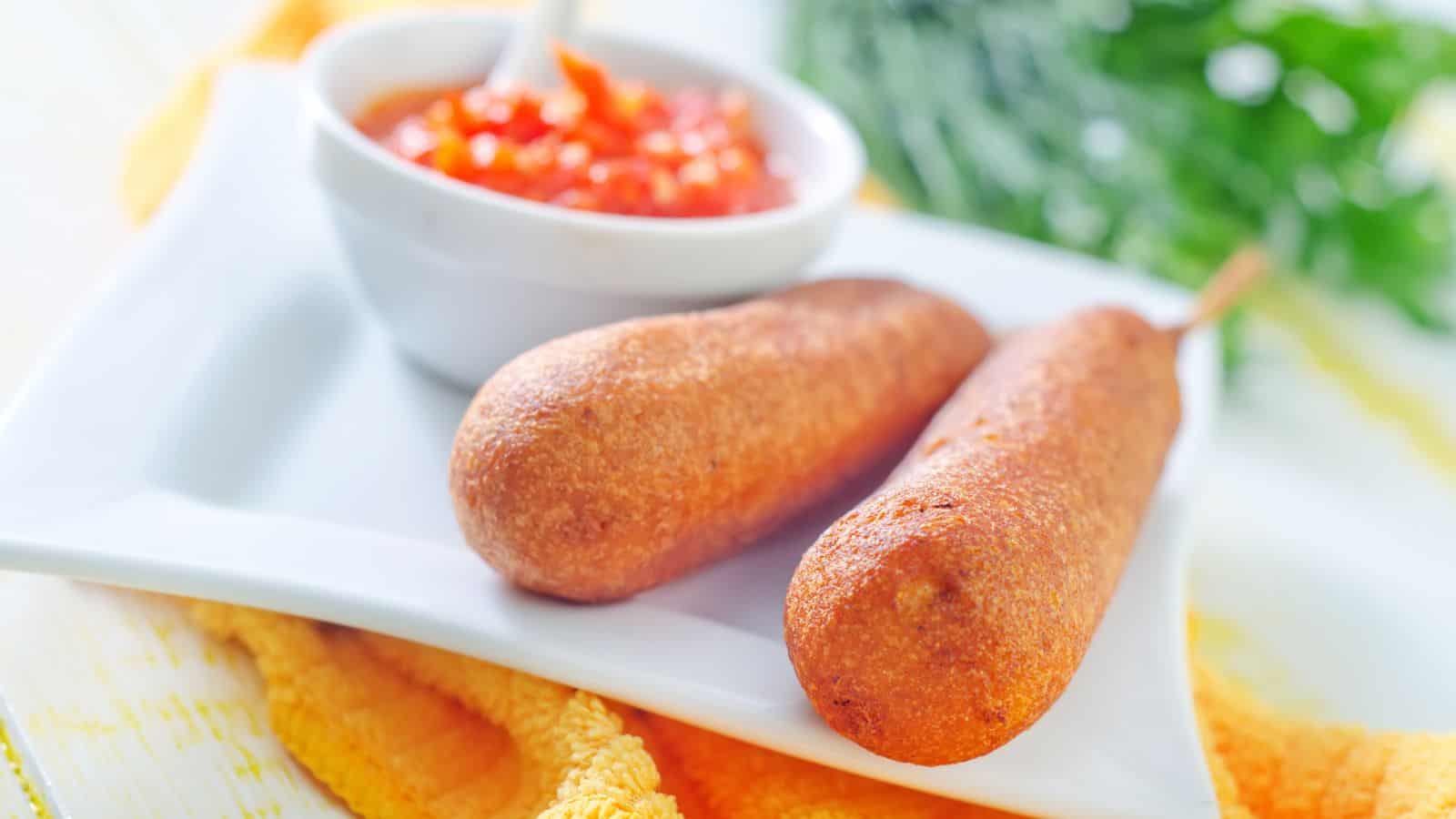 Two corn dogs on a white plate with a bowl of red dipping sauce and a spoon in the background, placed on a yellow cloth. Green leafy vegetables are partially visible in the background.