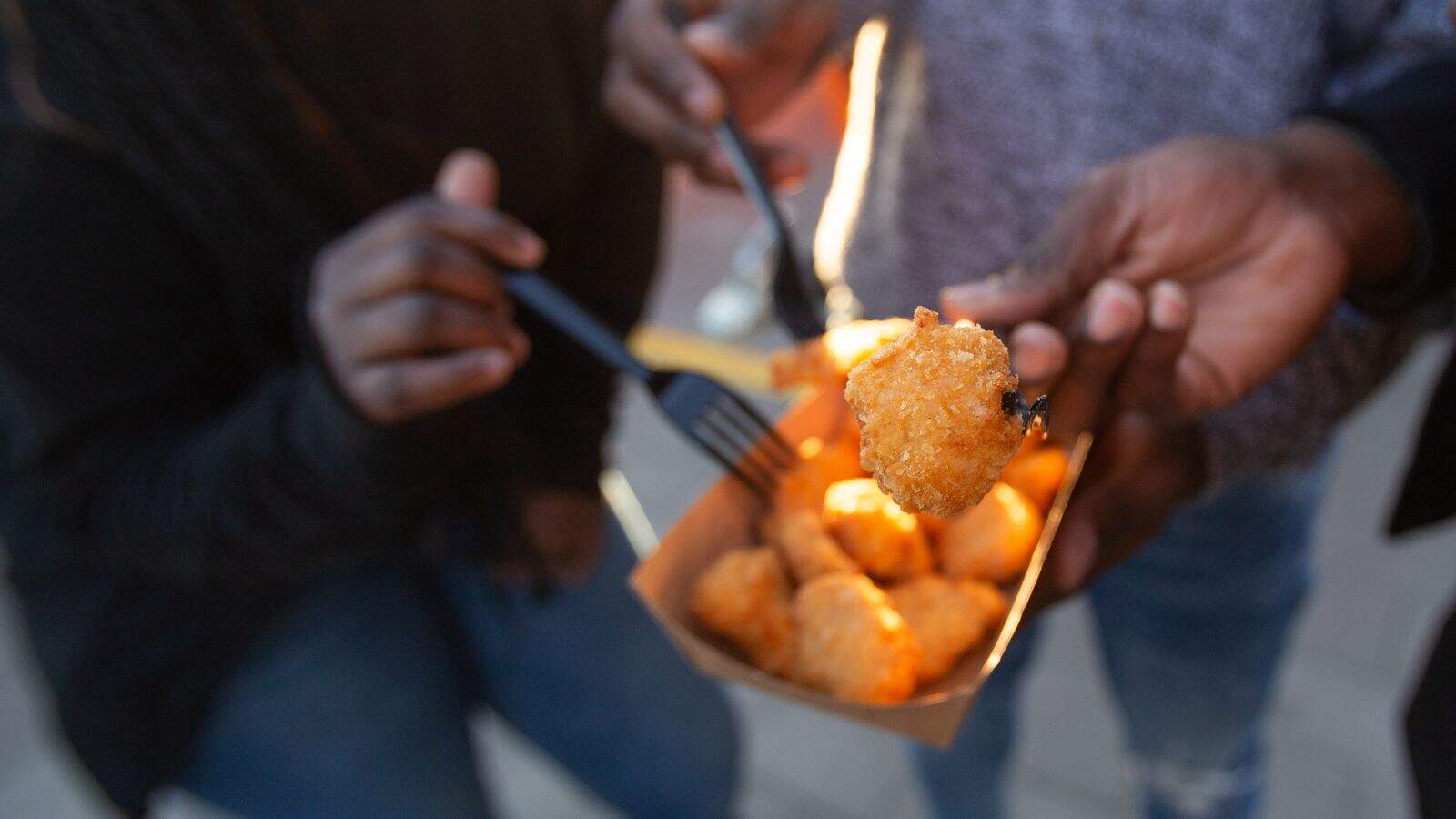 Three people hold forks and eat golden brown tater tots from a brown paper tray. The scene is outdoors and the focus is on the food, with hands holding the tater tots in the foreground.