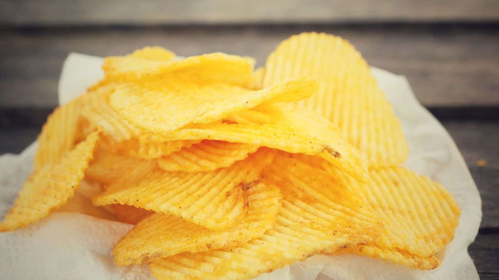 A pile of ridged potato chips, lightly seasoned, sits on a white paper napkin atop a wooden surface.