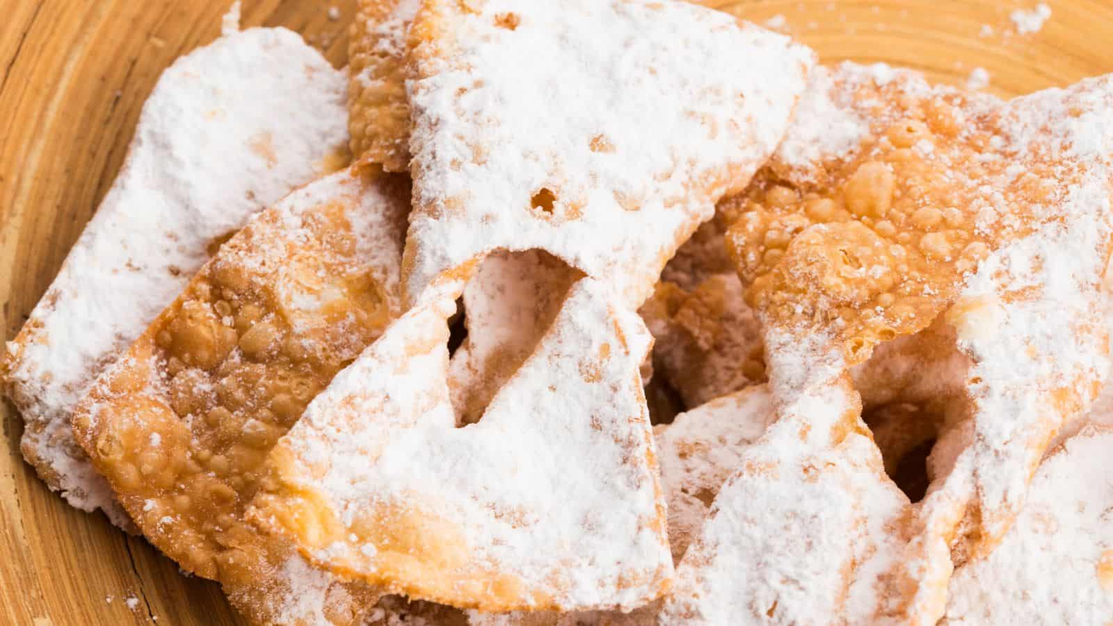 A close-up of several pieces of fried pastry dusted with powdered sugar, arranged on a wooden surface. The pastries have a bubbly texture and irregular shapes.