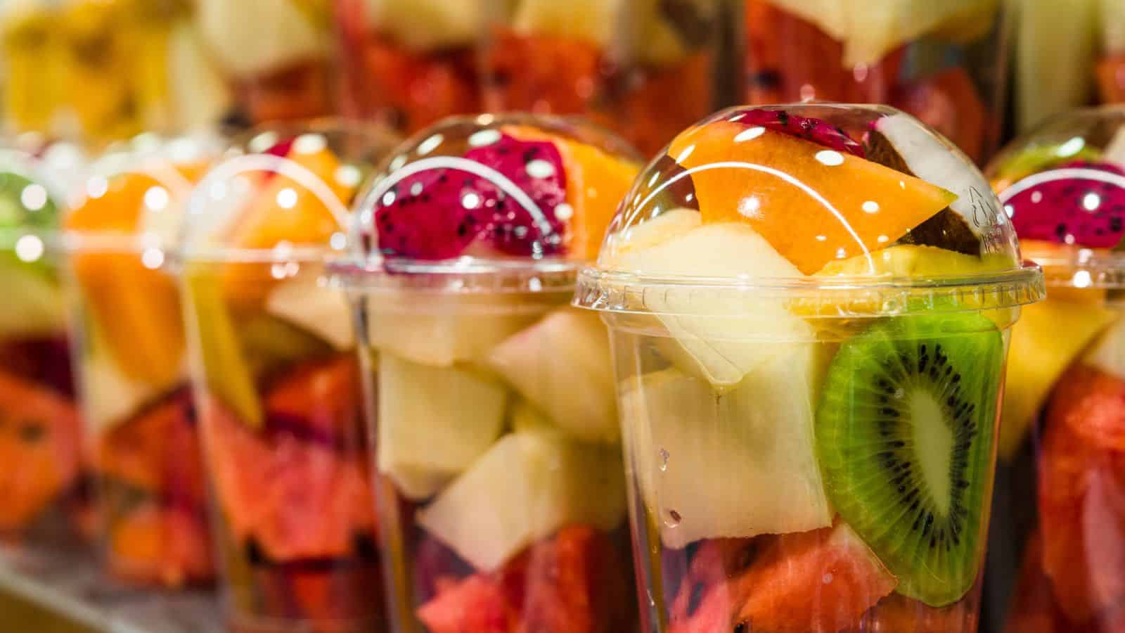 Clear plastic cups filled with assorted cut fruit, including watermelon, melon, kiwi, and dragon fruit, covered with dome lids, are displayed in rows.