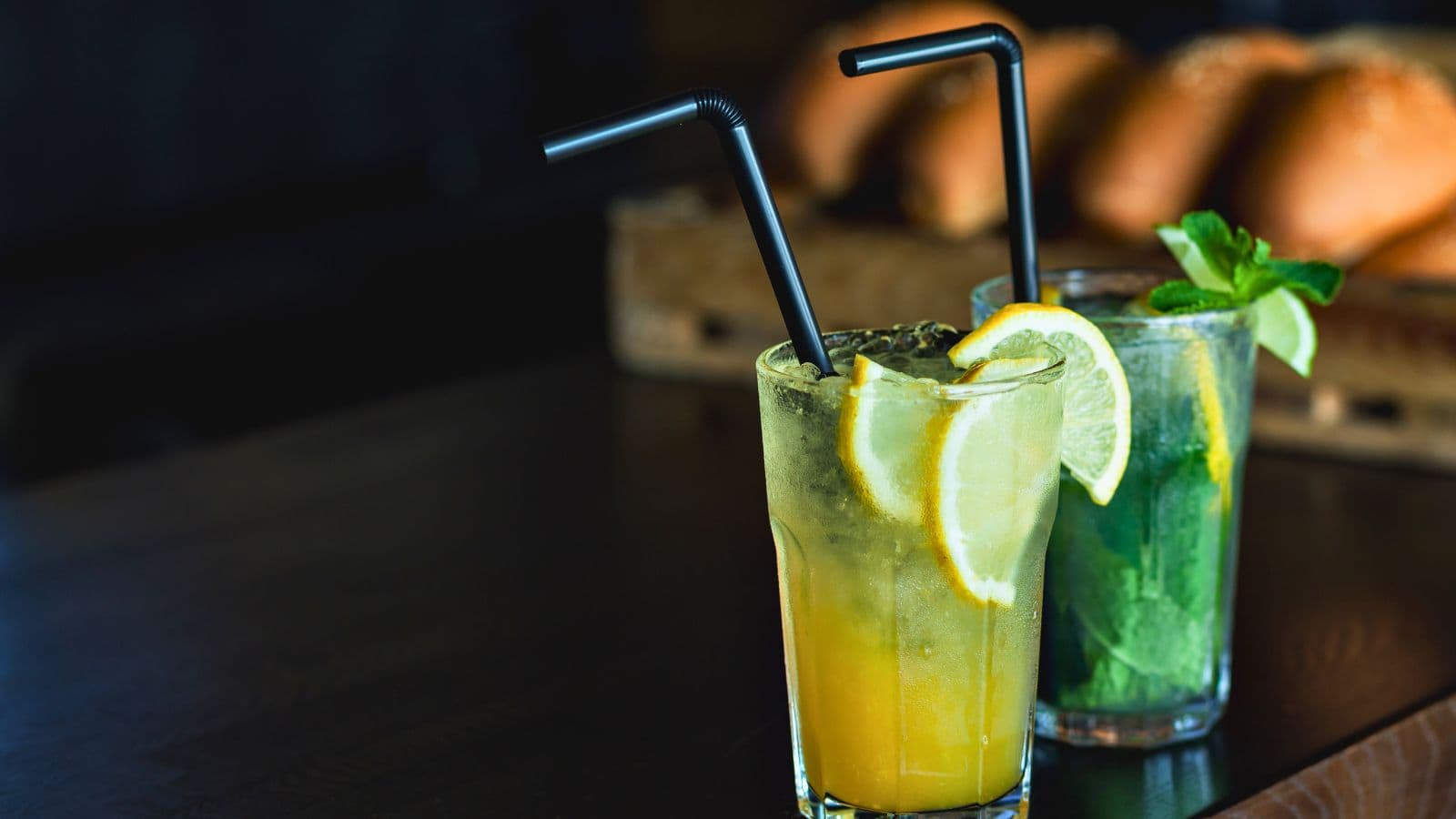 Two cocktails on a dark wooden table. The front glass contains a yellow drink with ice and lemon slices, garnished with a black straw. The back glass has a green drink with lime slices, mint, ice, and a black straw. Bread is in the background.