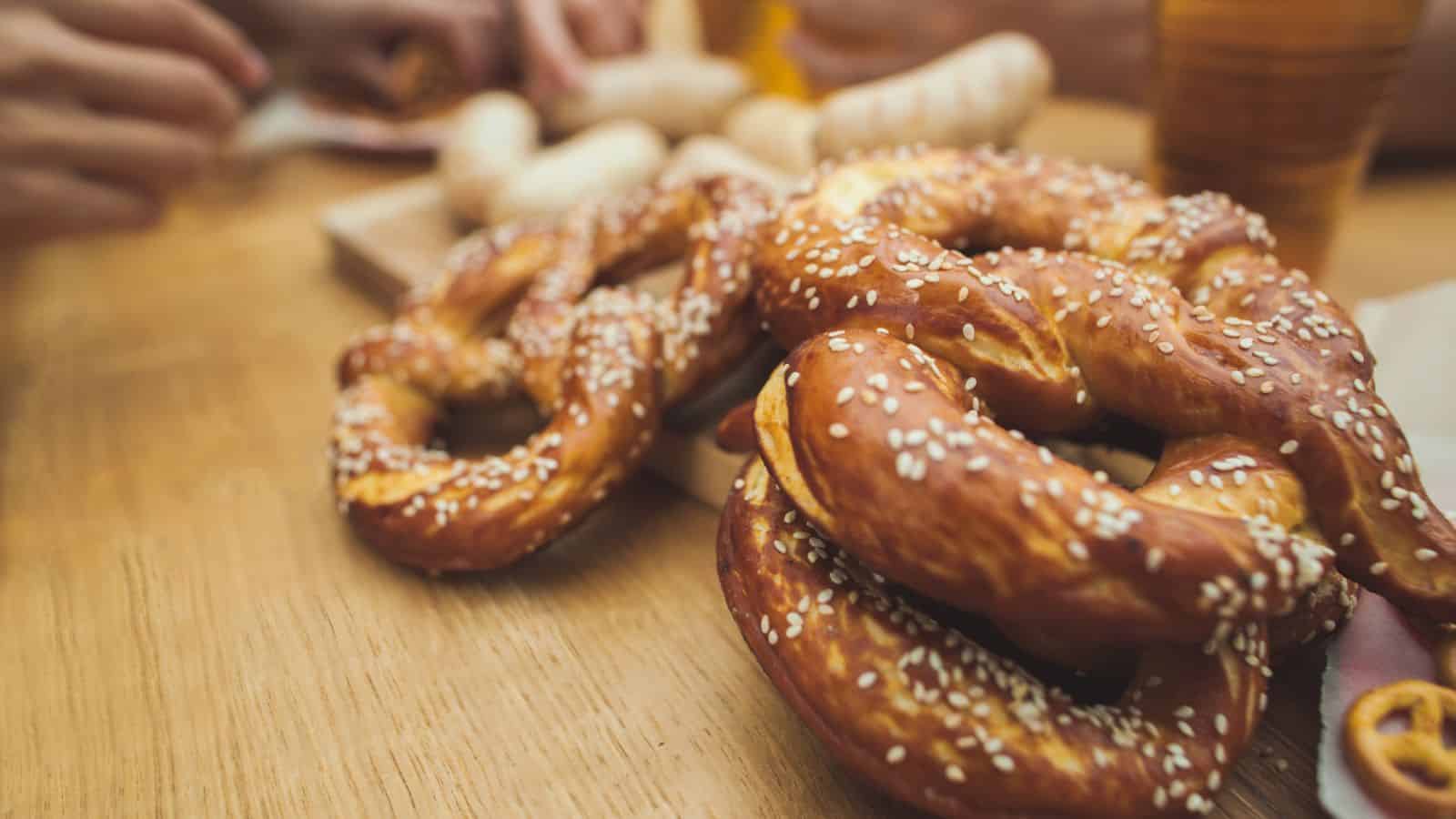 Close-up of salted pretzels with sesame seeds on a wooden table, with blurred hands, sausages, and beverages in the background.