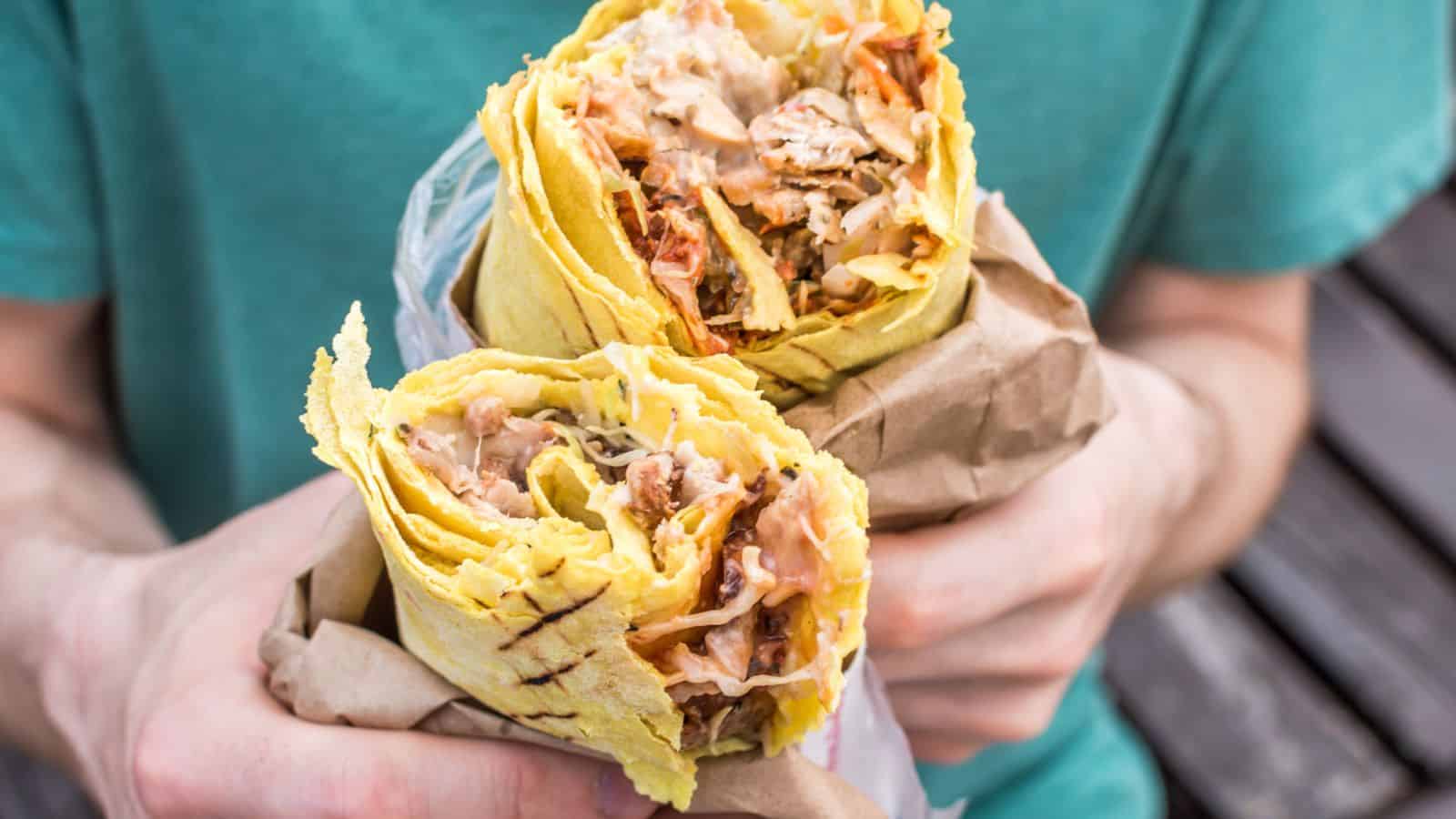 A person holding two halves of a shawarma wrap filled with chicken, vegetables, and sauce, wrapped in yellow flatbread and partially covered with brown paper. The person is wearing a green shirt.