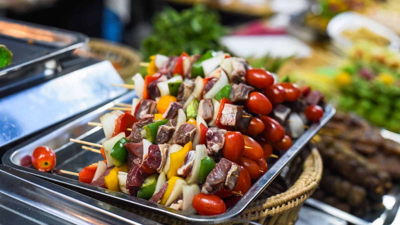 A metal tray holds skewers with raw beef, cherry tomatoes, yellow bell peppers, green bell peppers, onions, and mushrooms, arranged neatly and ready for grilling. Other food items are blurred in the background.
