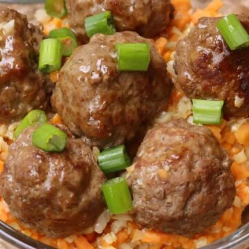 A glass bowl filled with six cooked meatballs garnished with chopped green onions, served on a bed of finely diced carrots and cauliflower. The bowl is placed on a woven surface.