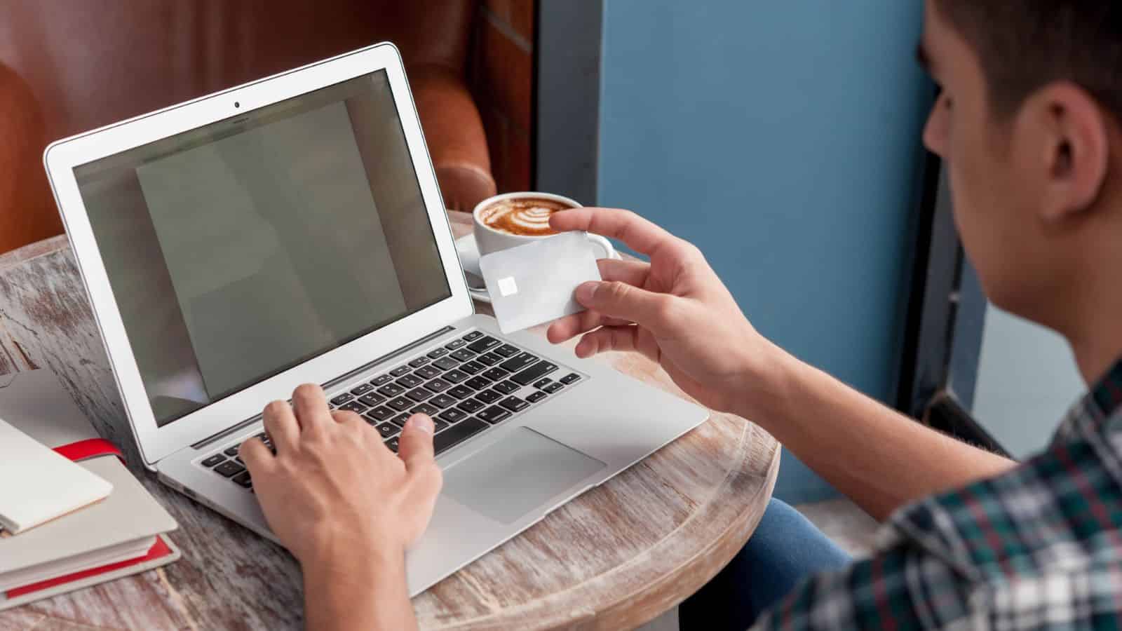 A person sits at a round wooden table using a laptop, holding a bank card in one hand. A cup of coffee, a notebook, and a pen are also on the table.