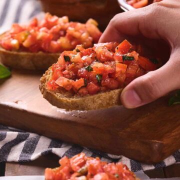 A hand holds a slice of toasted bread topped with diced tomatoes and herbs, with more pieces of bruschetta on a wooden board and a striped cloth beneath.