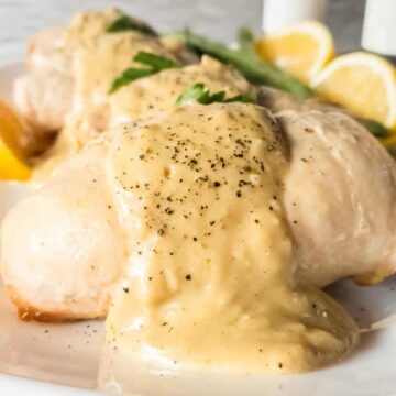 Three pieces of cooked chicken breast covered in a creamy sauce and sprinkled with black pepper are served on a white plate, garnished with lemon wedges and fresh parsley.