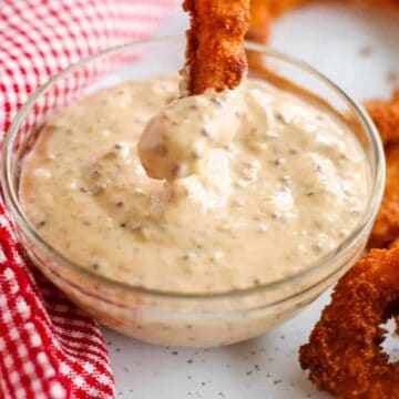 A clear glass bowl of creamy, speckled dipping sauce sits on a surface next to a red and white checkered cloth. A breaded onion ring is being dipped into the sauce, with other onion rings nearby.