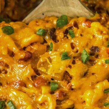 Close-up of cheesy macaroni mixed with beans, vegetables, and topped with chopped green onions, with a wooden spoon partially visible.