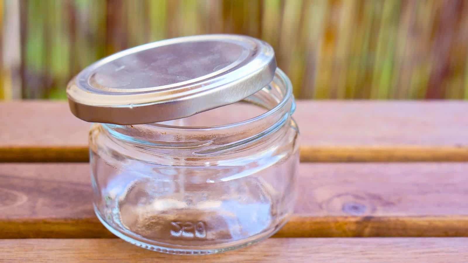 A small empty glass jar with a loose metal lid sits on a wooden surface, subtly hinting at potential canning dangers if not properly sealed.