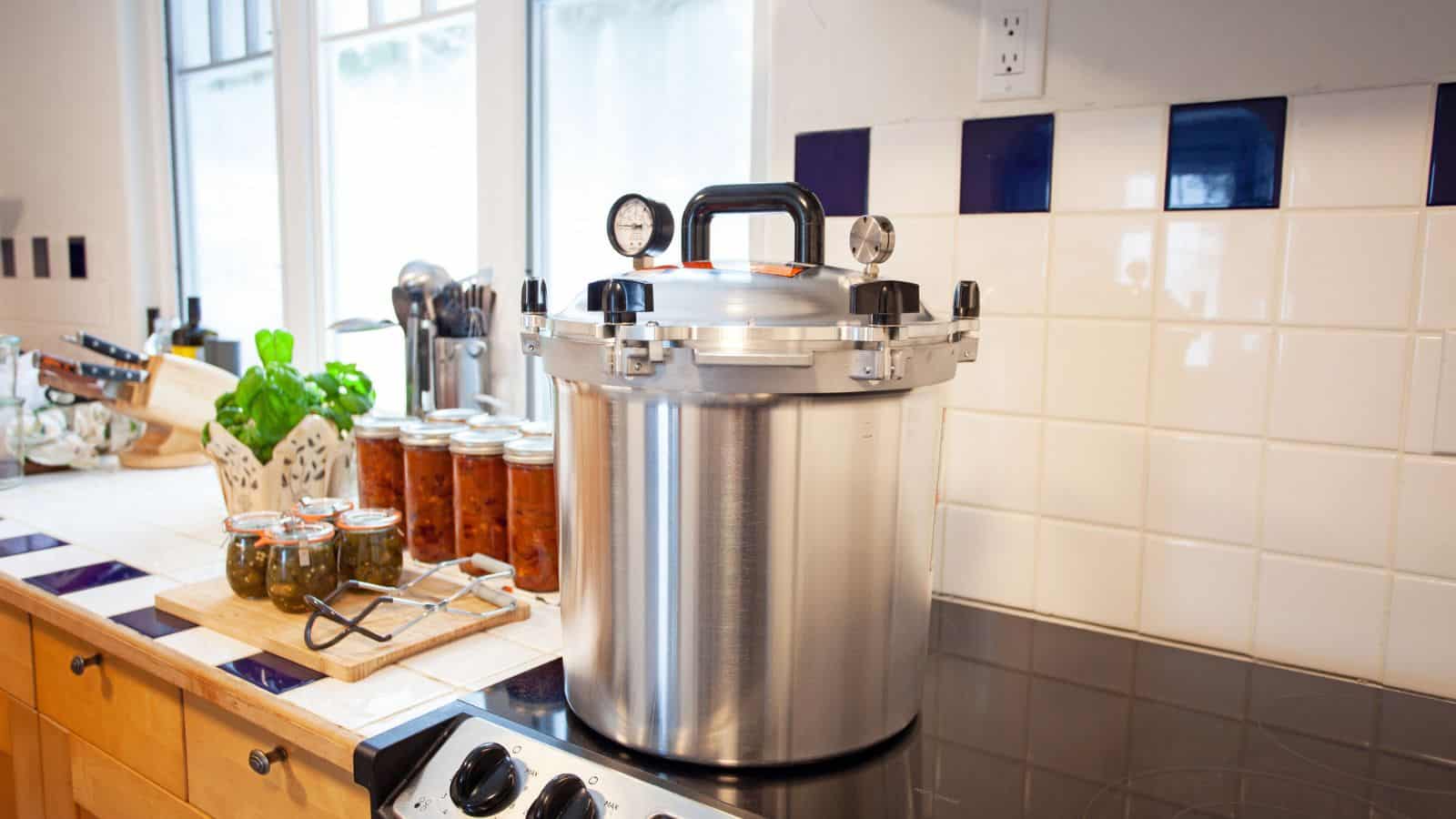 A large metal pressure canner sits on a kitchen stove near jars of preserved food and kitchen utensils, serving as a reminder to always be mindful of canning dangers during the preservation process.