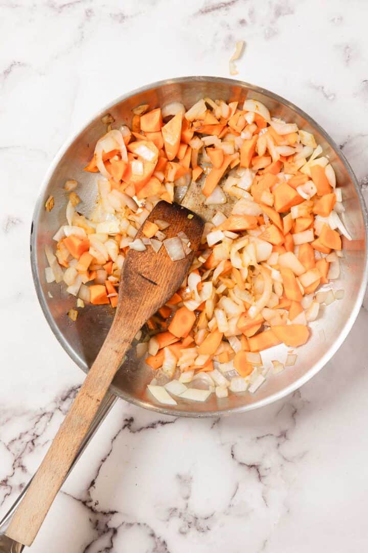 Chopped onions and carrots being sautéed in a pan with a wooden spoon on a marble surface, perfect as the flavorful base for Chicken Borscht.