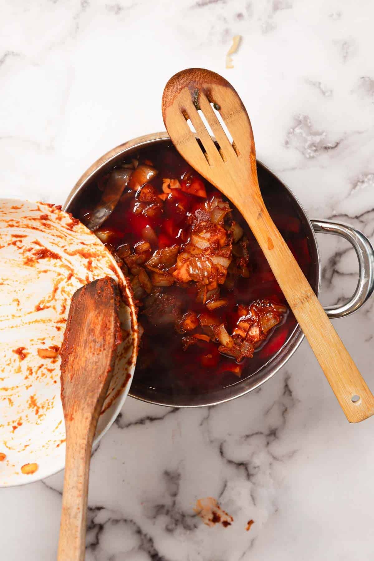 A pot of Chicken Borscht stew with a wooden slotted spoon and a messy bowl with a wooden spoon rest on a marble countertop.