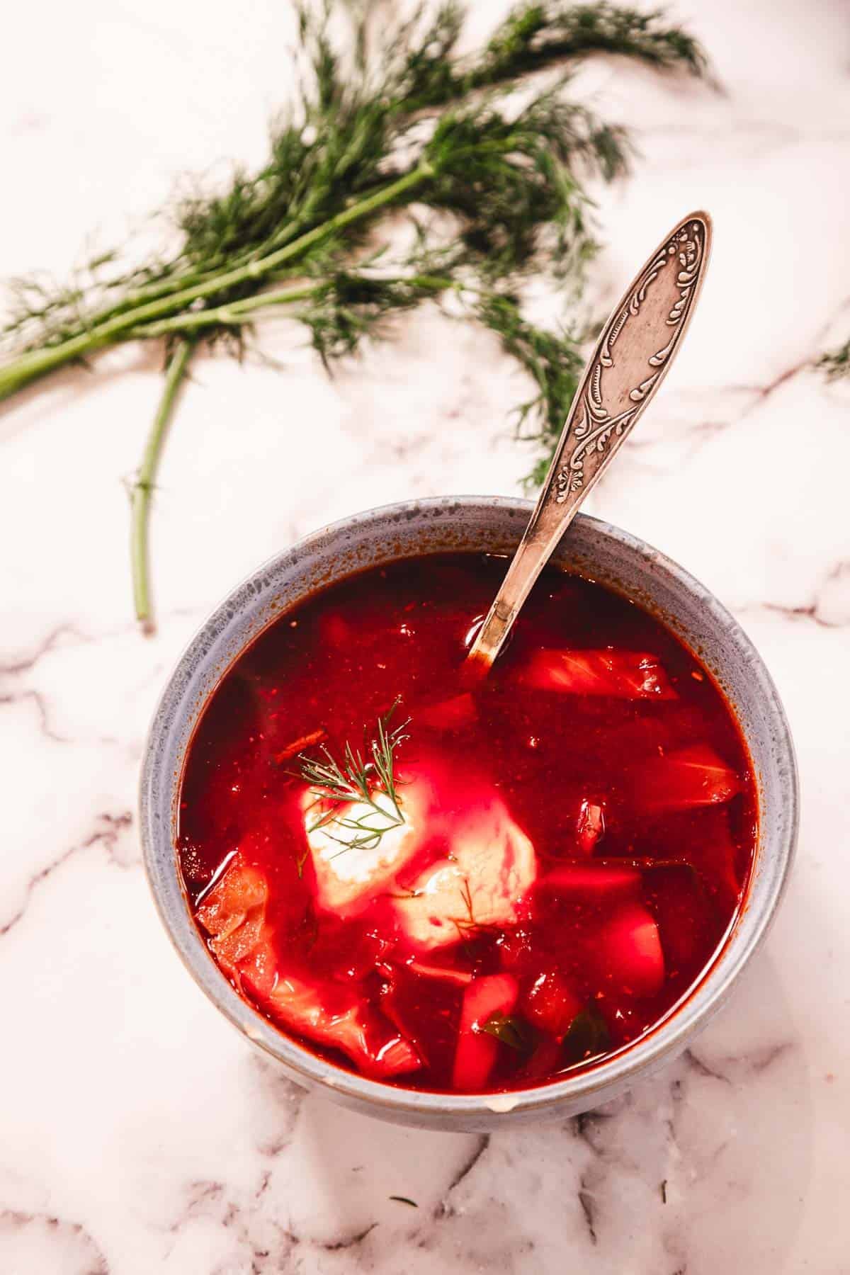 A bowl of red soup with a dollop of sour cream, garnished with dill, and a spoon, on a marble surface.