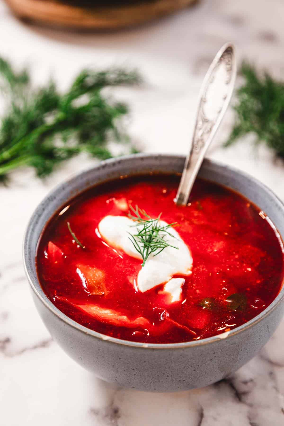 A bowl of red soup with sour cream and dill garnish, with a spoon in the bowl on a marble surface.