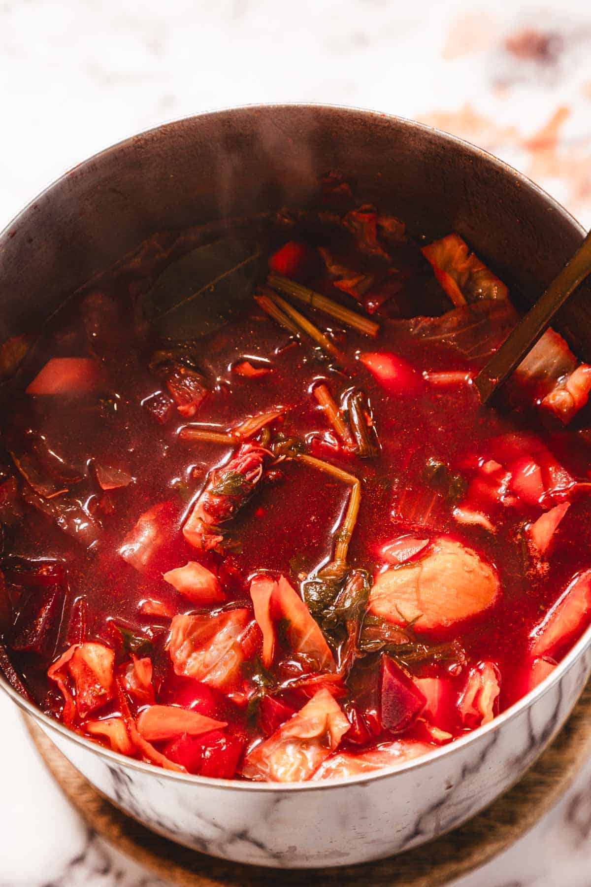 A pot of vibrant red borscht soup filled with vegetables and leafy greens, steaming hot.