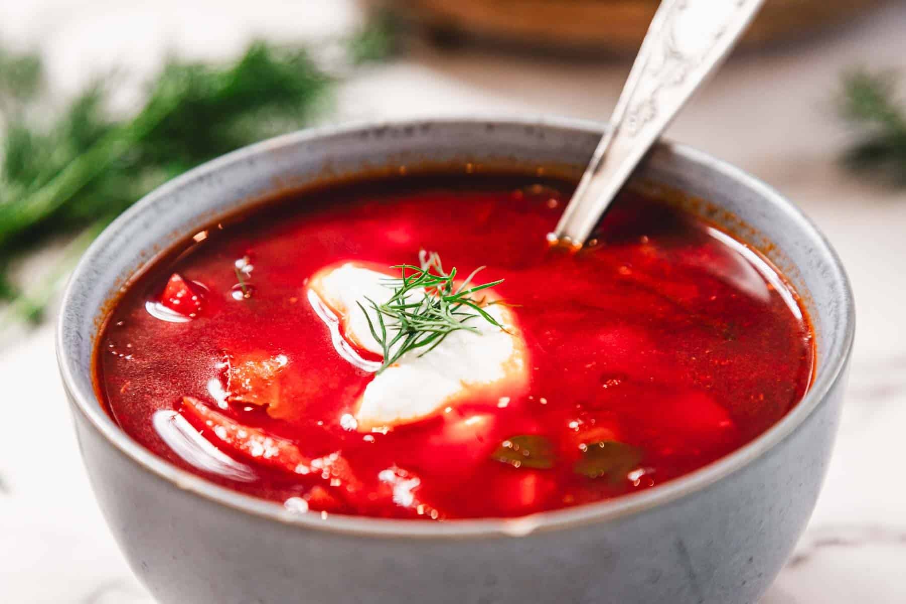 A bowl of red borscht soup with a dollop of sour cream and dill, and a spoon inside.