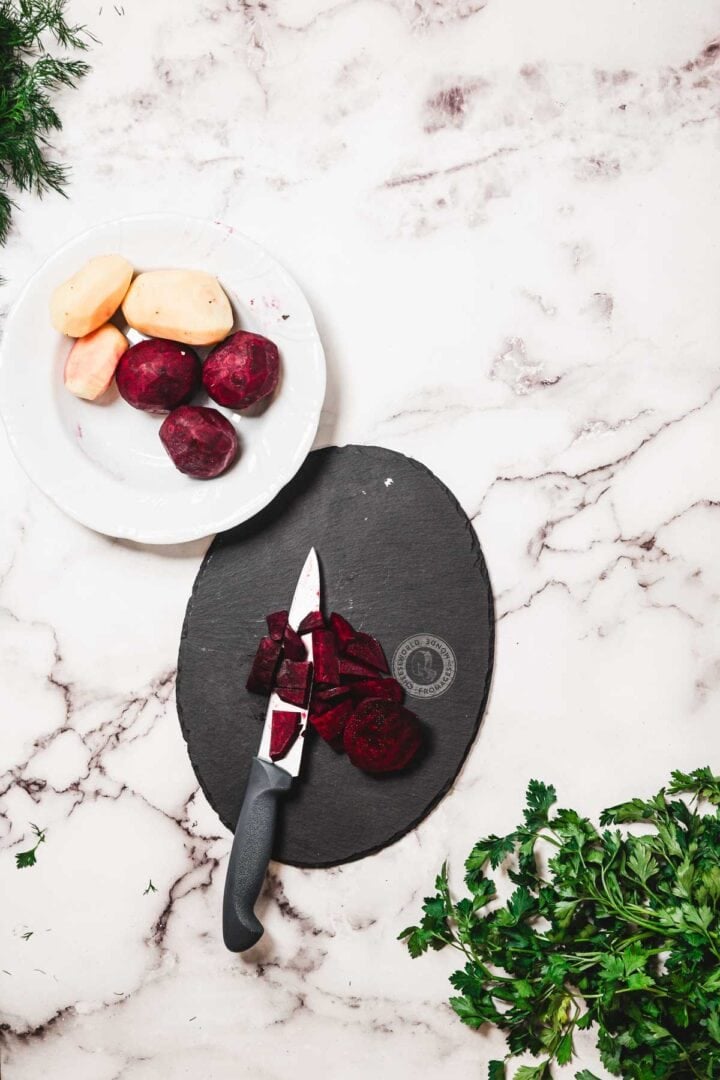 Sliced beets and a knife on a slate board, with whole beets and potatoes on a plate, herbs on the side—perfect ingredients for preparing hearty Chicken Borscht.