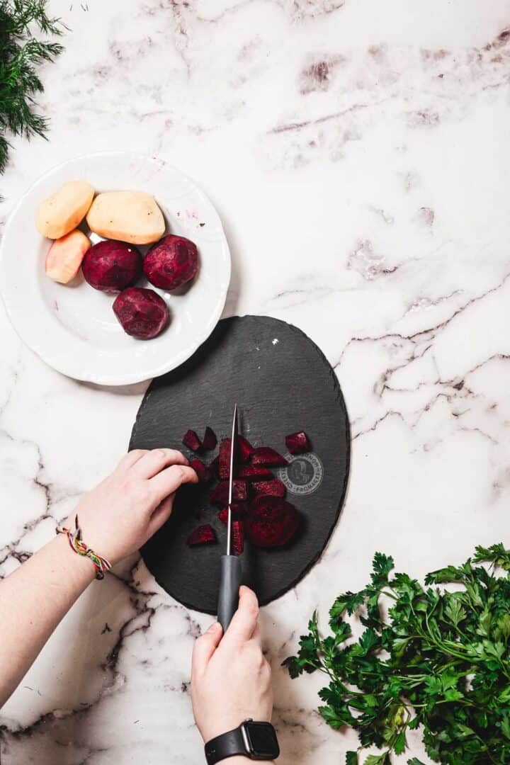 Hands slicing beets on a black cutting board, prepping veggies for Chicken Borscht, with potatoes on a plate and parsley on a marble counter.