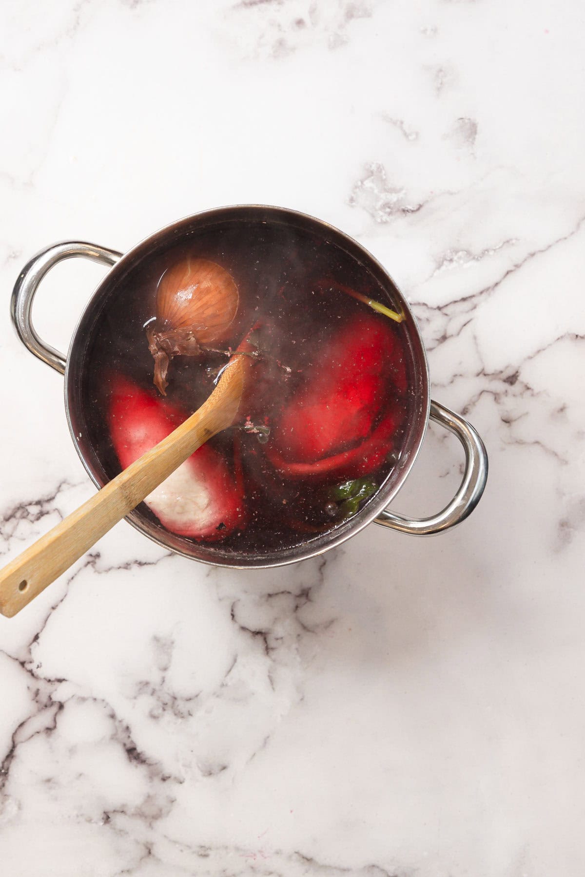 A pot of simmering Chicken Borscht with tomatoes, onion, herbs, and a wooden spoon sits on a marble surface.